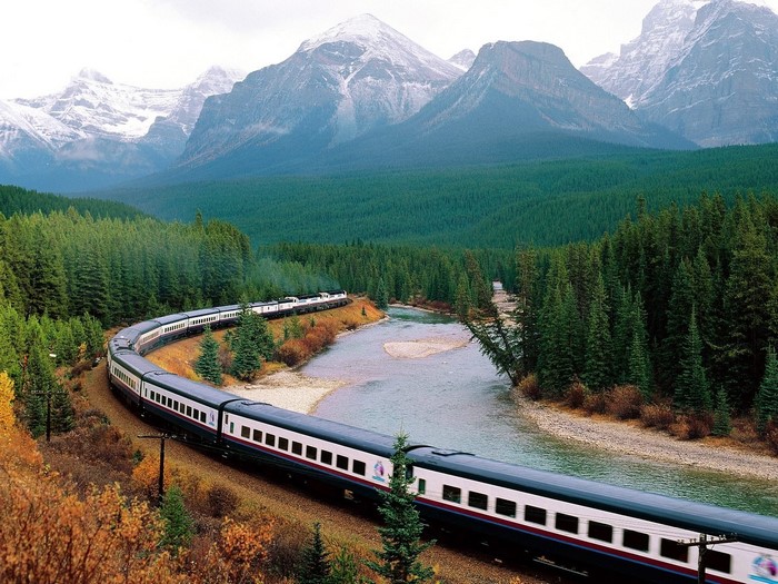 Train traveling through a forest with mountains and a river in Canada