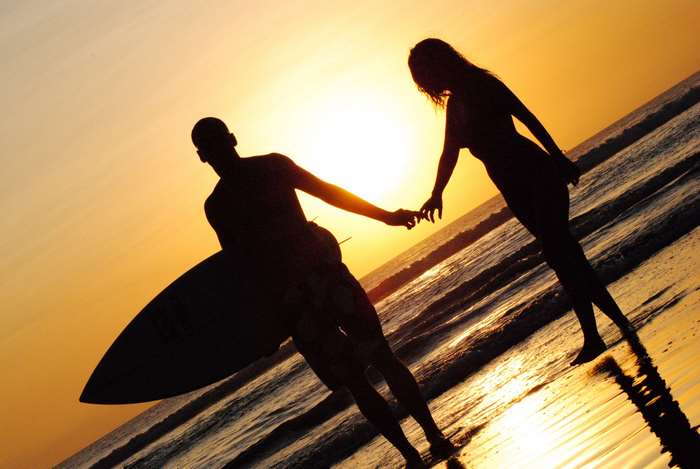 Couple holding hands at beach sunset with surfboard