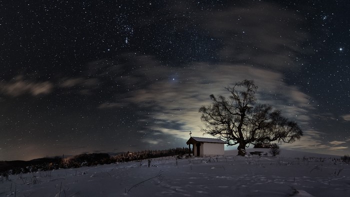 Winter night sky over a snowy landscape with a chapel and tree