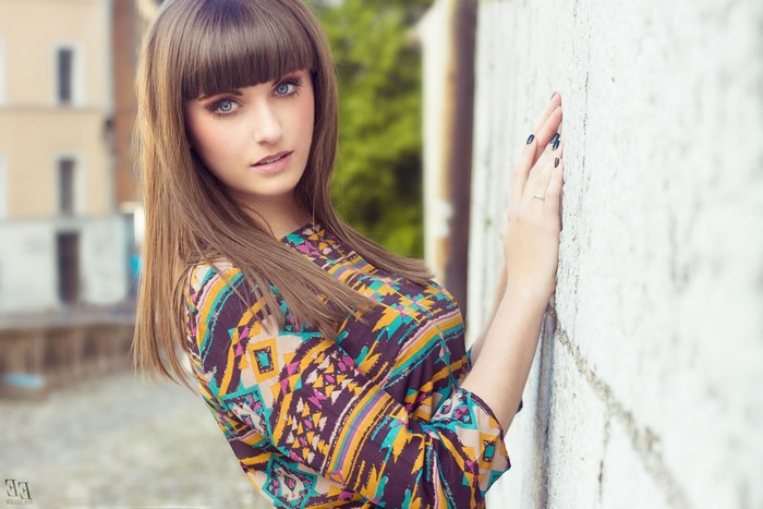 Woman with long brown hair and bangs leaning against a wall