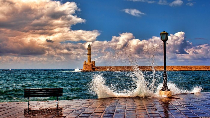 Waves crashing near a lighthouse and lamp post in Crete