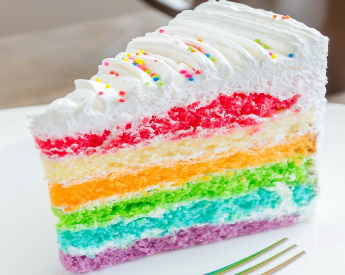 Slice of rainbow layer cake with white frosting and sprinkles