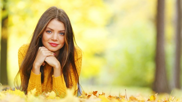 Woman with long brown hair wearing a yellow sweater lying in fall leaves