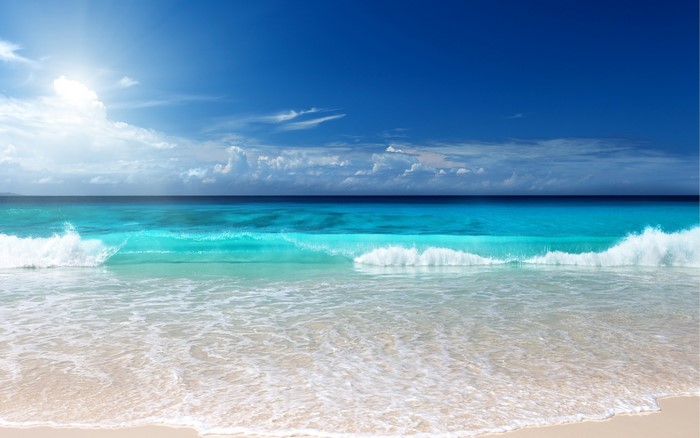 Turquoise ocean waves crash on a sandy beach under a blue sky with clouds