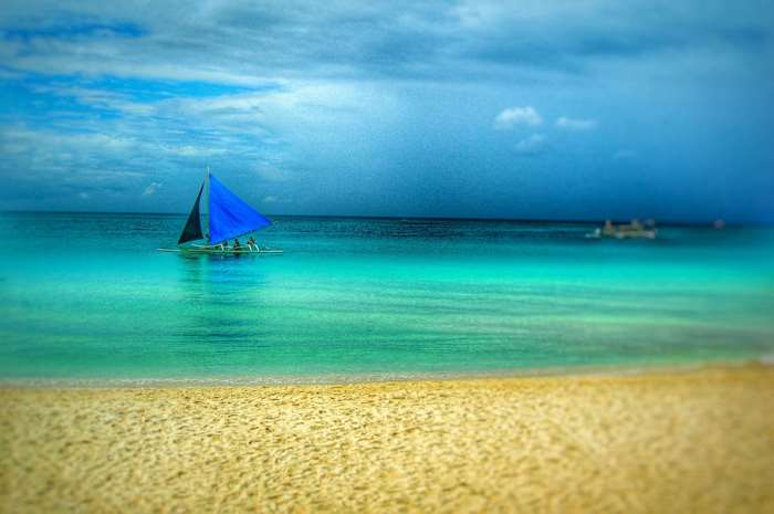 Sailboat with blue sail on turquoise water near sandy beach