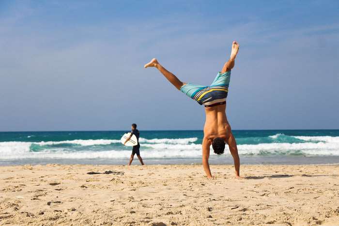 Man performs handstand on beach with surfboarder in background