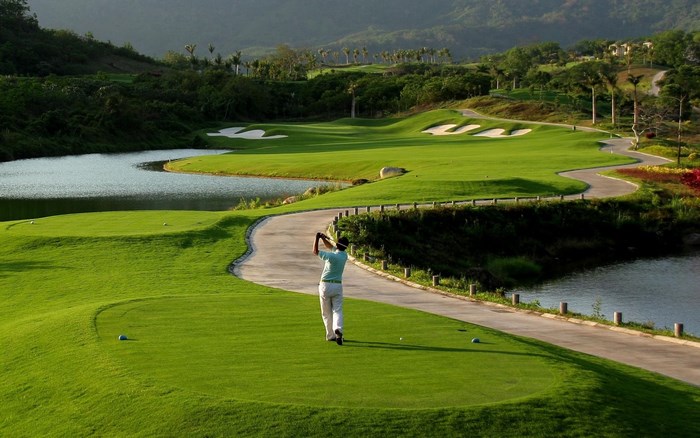 Golfer swings club on a scenic golf course with water hazards and sand traps.