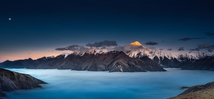 Snowy mountain peaks above clouds at sunset with moon
