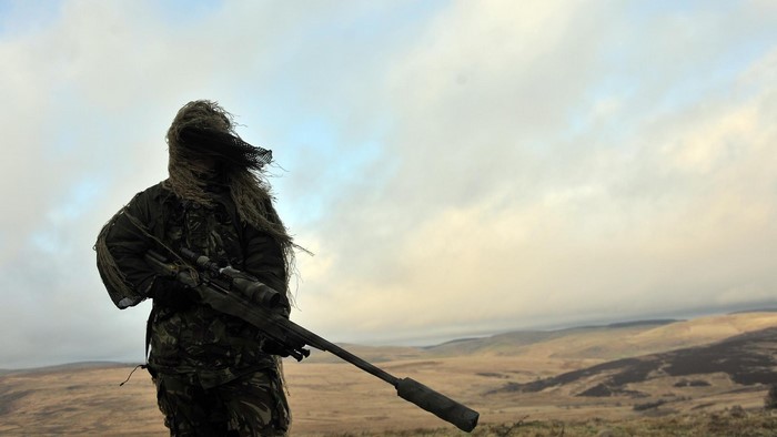 Soldier in ghillie suit with sniper rifle in hilly terrain