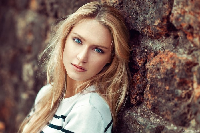 Woman with blonde hair and blue eyes leaning against a rock wall