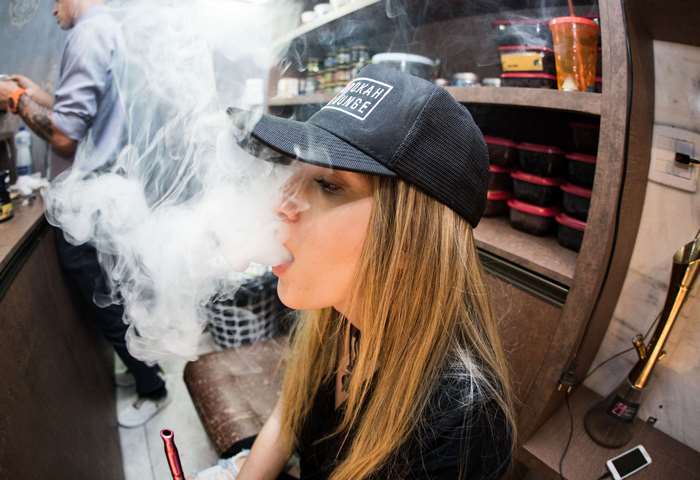 Woman wearing a Hookah Lounge cap exhales smoke