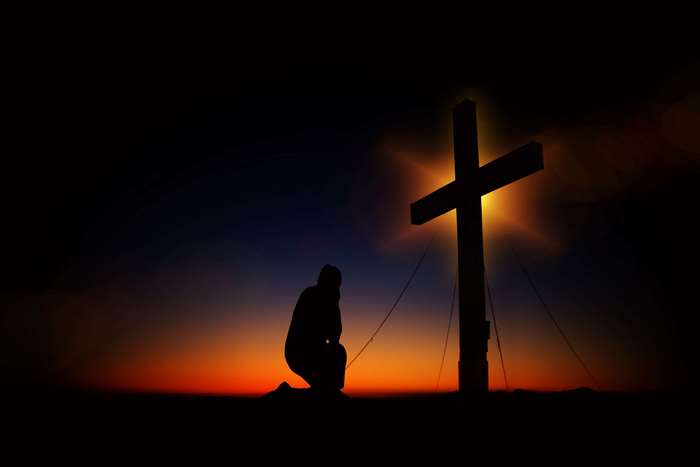 Silhouette of a person kneeling before a cross at sunset