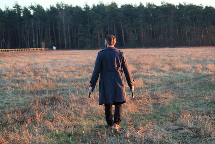 Man in coat holding two guns in a field