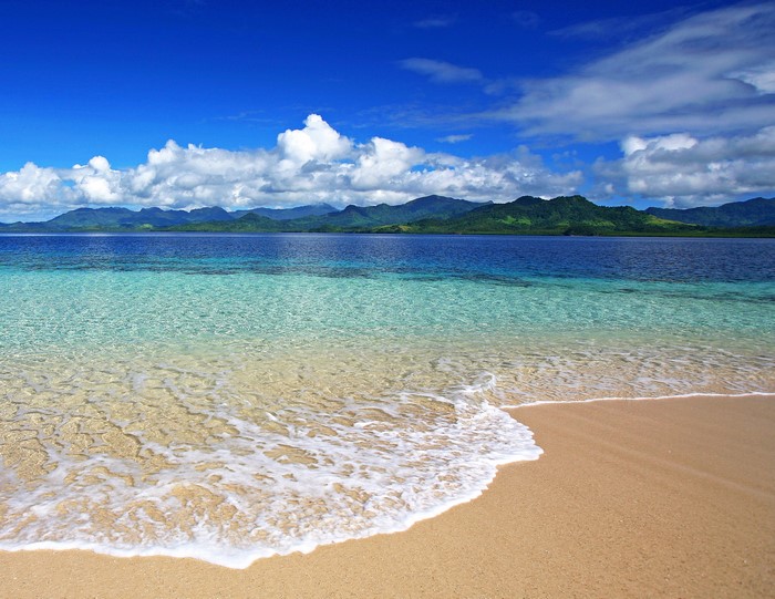 Crystal clear turquoise water laps onto a sandy beach with green mountains in the distance under a blue sky with clouds.