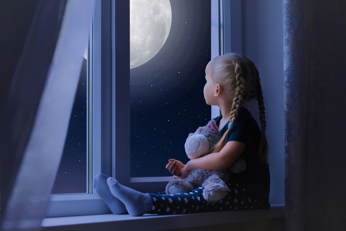 Little girl with pigtails looking at the moon through a window
