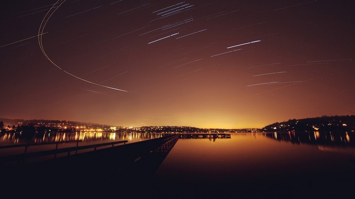 Night sky with star trails over city lights and a pier