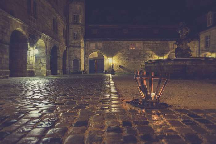 Cobblestone courtyard with arches and a torch at night