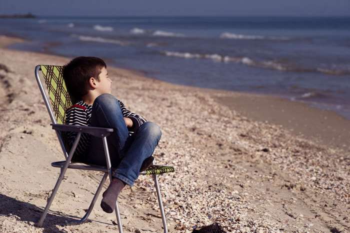 Boy sitting on a chair at the beach looking at the ocean