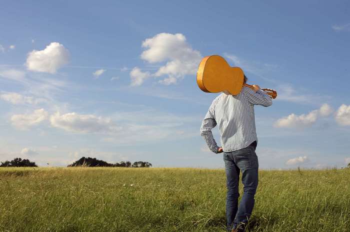 Man carrying a guitar in a grassy field under a blue sky