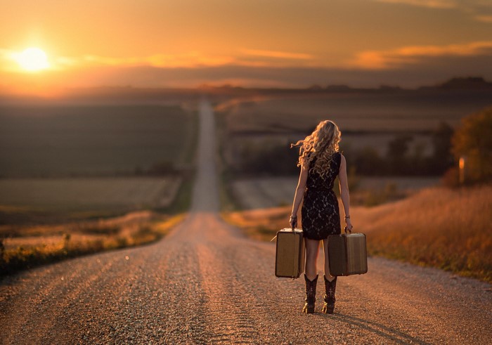 Woman with suitcases walking on a rural road at sunset