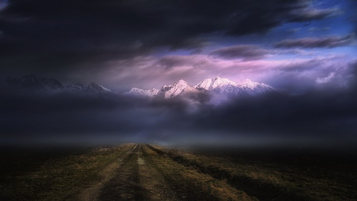 Dirt road leading to snowy mountains under dark clouds