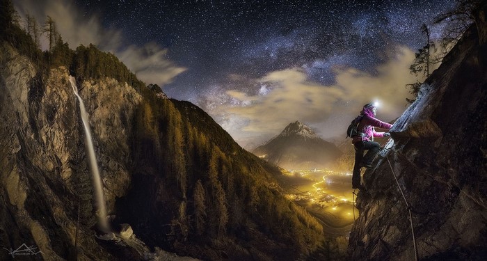 Woman rock climbing at night with waterfall and village below