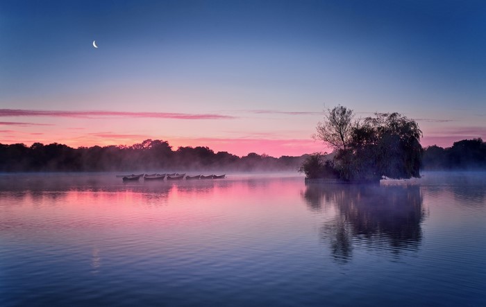 Calm lake with boats at sunrise and crescent moon