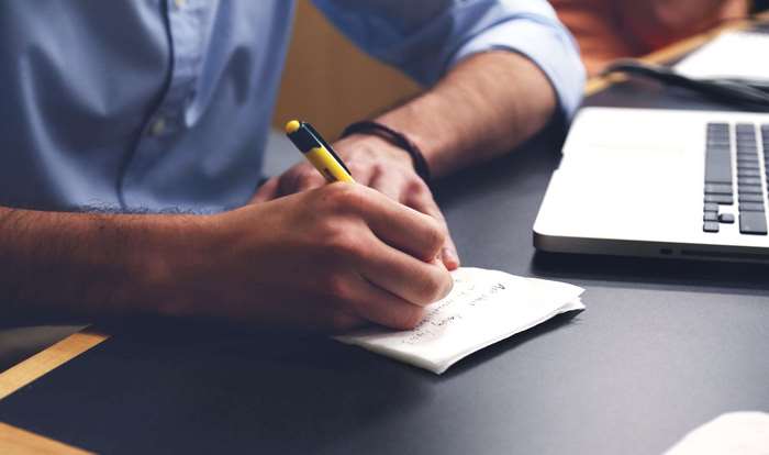 Person writing notes on paper with a pen next to a laptop