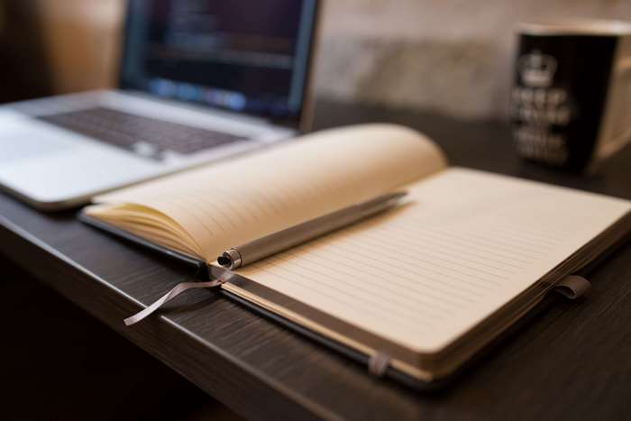 Notebook and pen on a desk with a laptop and mug