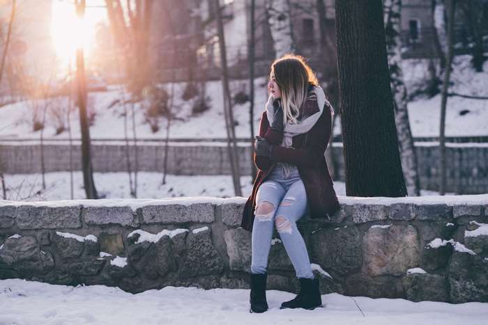 Woman in winter coat and ripped jeans sitting on a snowy wall