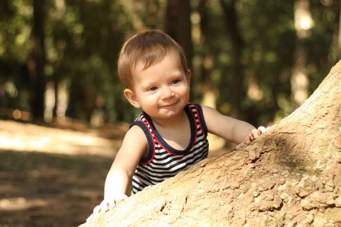 Baby boy smiling near a tree trunk in a park