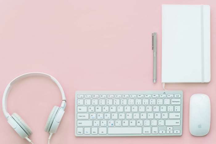 White headphones, keyboard, mouse, notebook, and pen on pink background
