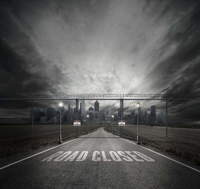 Road closed to a fenced-off city under stormy skies