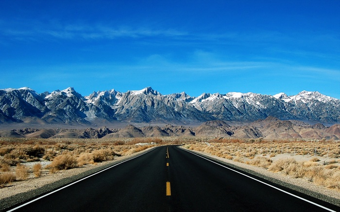 Desert road leading to snow-capped mountains under a blue sky