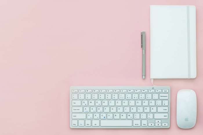 White keyboard, mouse, notebook, and pen on pink background