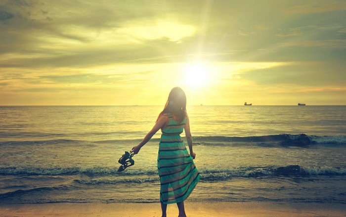 Girl in dress holding shoes on beach at sunset