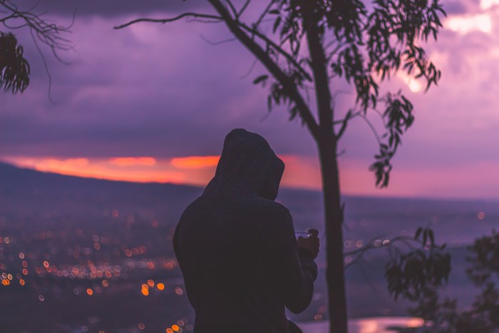 Person in hoodie with phone overlooking cityscape at sunset
