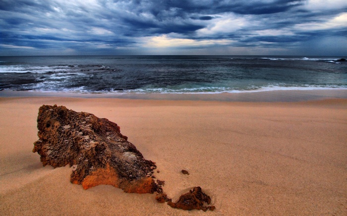 A large rock on a sandy beach with stormy ocean waves.