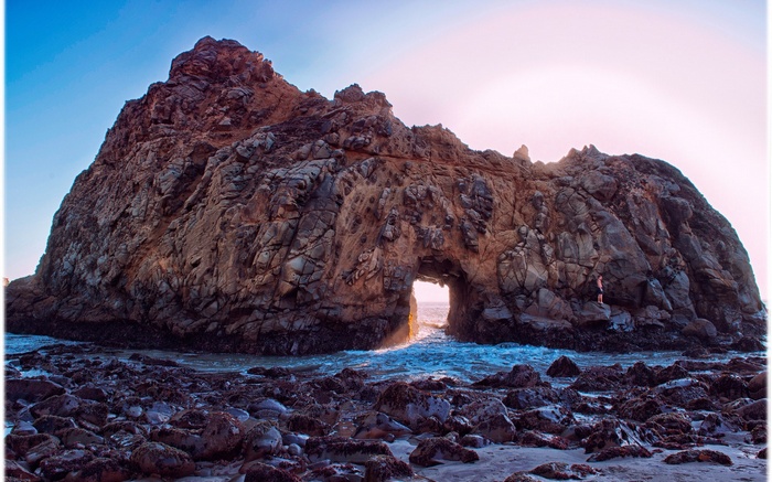 Man standing on rocks near a sea arch at sunset