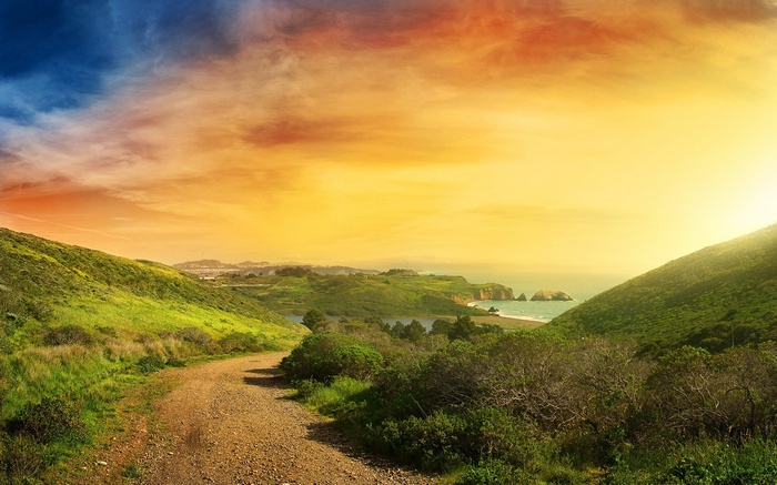 Dirt path through green hills towards the ocean at sunset