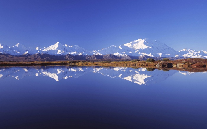 Snow-capped mountains reflected in a calm lake under a blue sky
