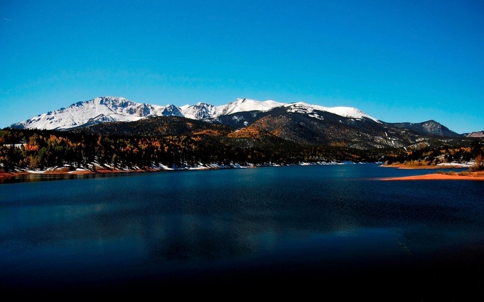 Snow-capped mountains overlook a dark blue lake and autumn forest