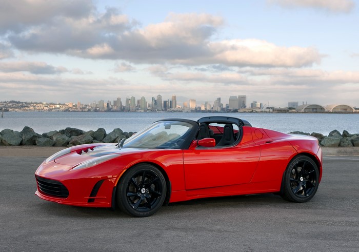 Red Tesla Roadster parked by the water with city skyline