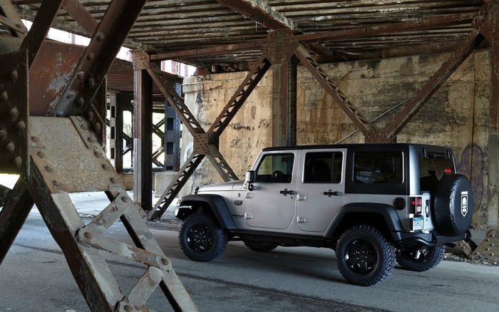 Silver Jeep Wrangler parked under a rusty bridge