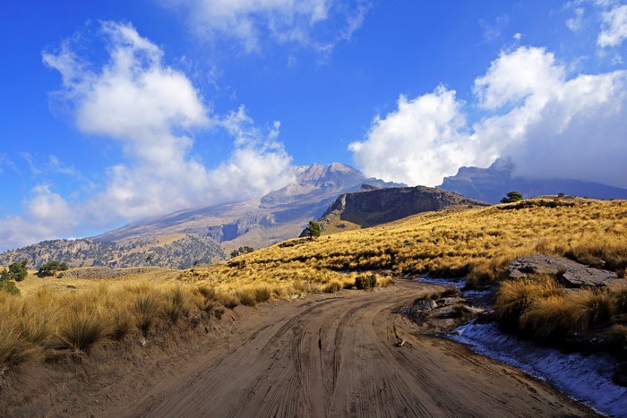 Dirt road leading to a mountain range under a blue sky with clouds
