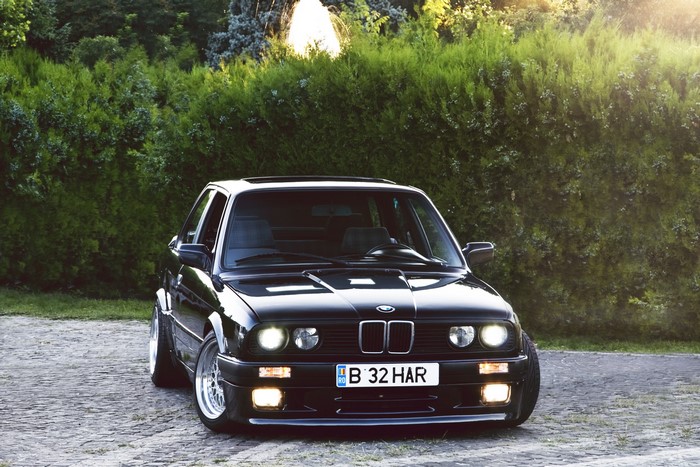 Black BMW E30 parked on cobblestone with trees in background