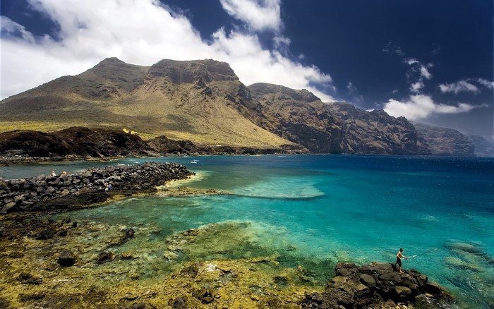 Man fishing by rocky coast with mountains