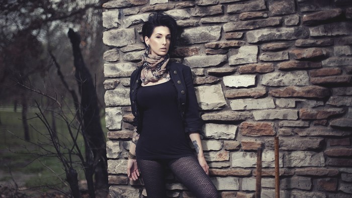 Woman in black dress and patterned pantyhose leaning against stone wall