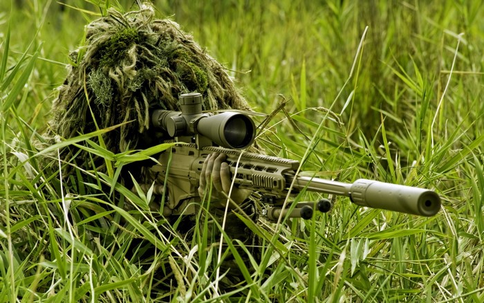 Soldier in ghillie suit aiming sniper rifle in grass