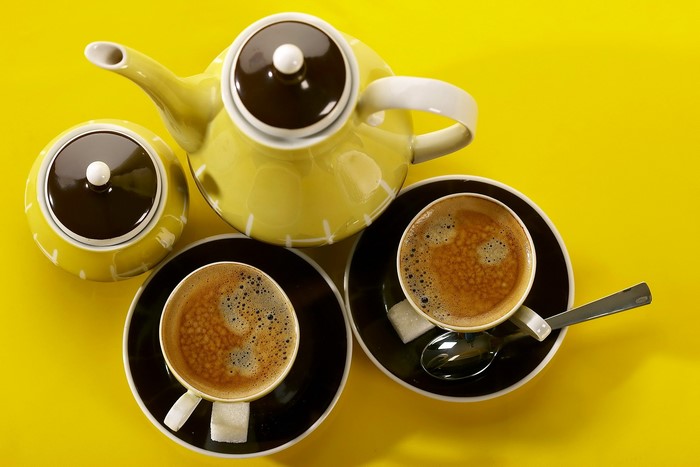 Yellow teapot, sugar bowl, and two cups of coffee on yellow background